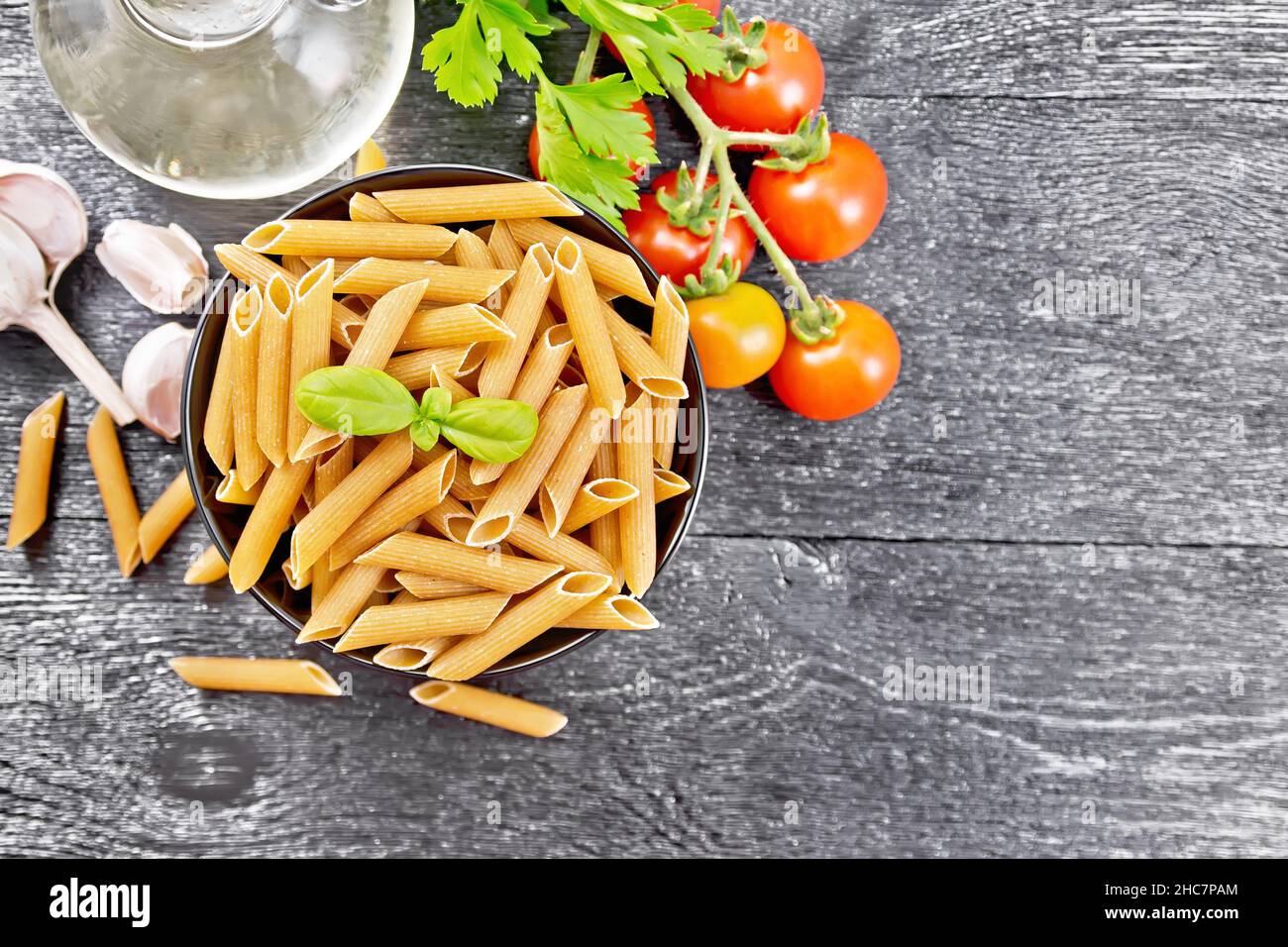 Whole grain flour penne pasta in a bowl, tomatoes, garlic, vegetable oil in a decanter and parsley on wooden board background from above Stock Photo