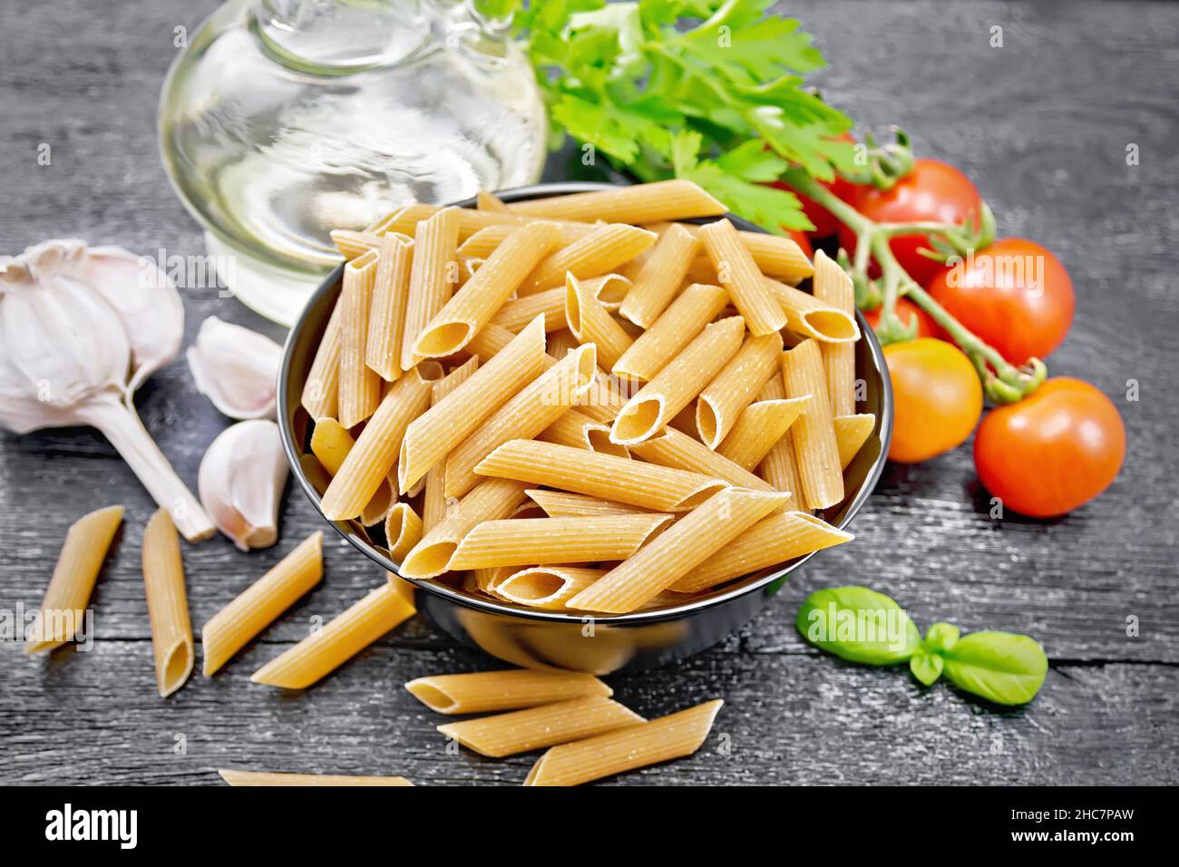 Whole grain flour penne pasta in a bowl, tomatoes, garlic, vegetable oil in a decanter and parsley on black wooden board background Stock Photo