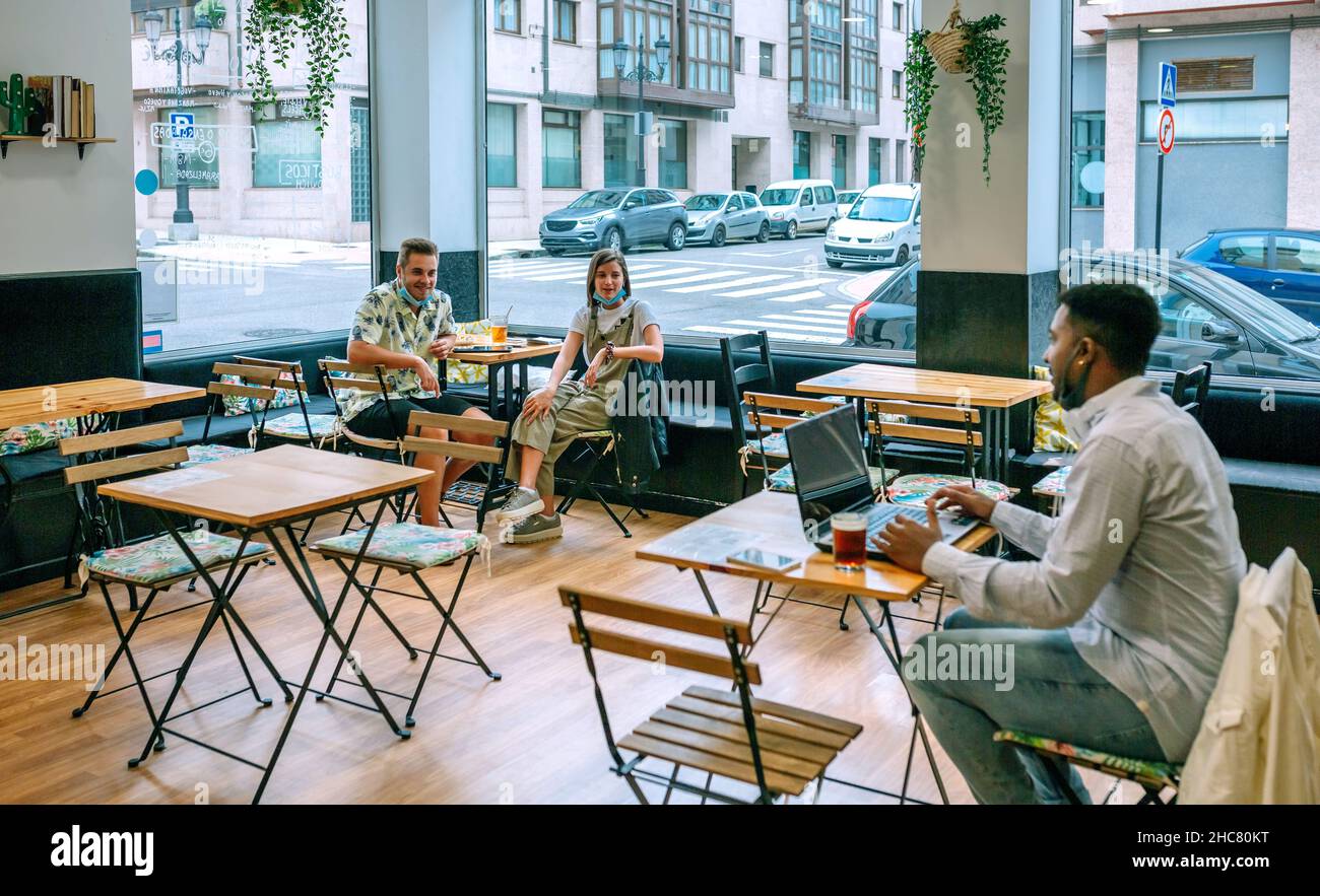 Young man teleworking in coffee shop talking to friends maintaining social distance Stock Photo