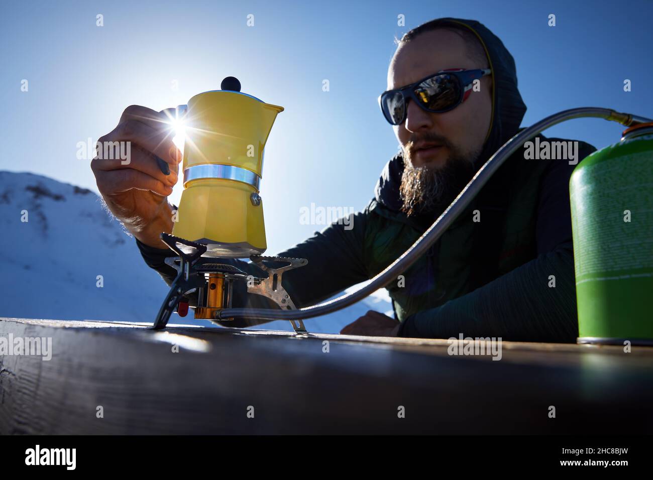 Bearded man hiker making coffee from Yellow Moka mocha pot on gas stove outdoors in the snow winter mountains. Old style coffee vintage pot outdoor ca Stock Photo