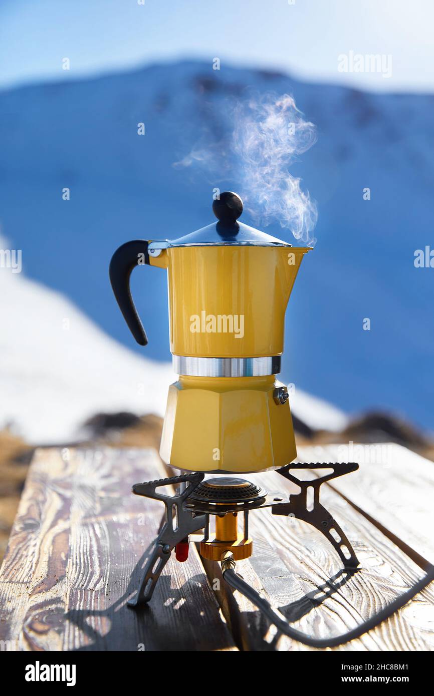 Yellow moka pot coffee with steam at snow mountain camping. Morning picnic, person cooking hot drink for breakfast in nature Stock Photo