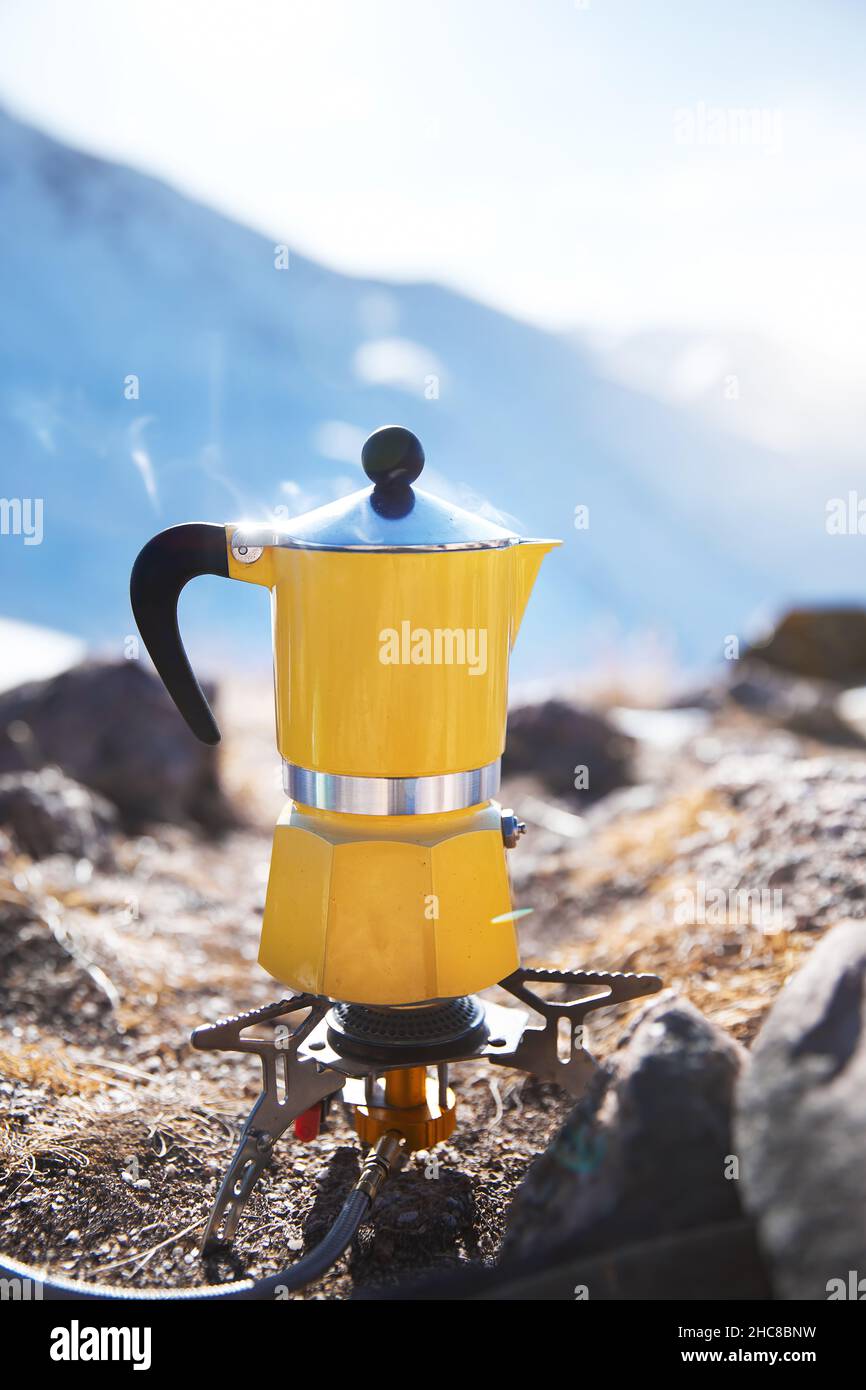 Yellow moka pot coffee with steam at snow mountain camping. Morning picnic, person cooking hot drink for breakfast in nature. Vertical shot Stock Photo