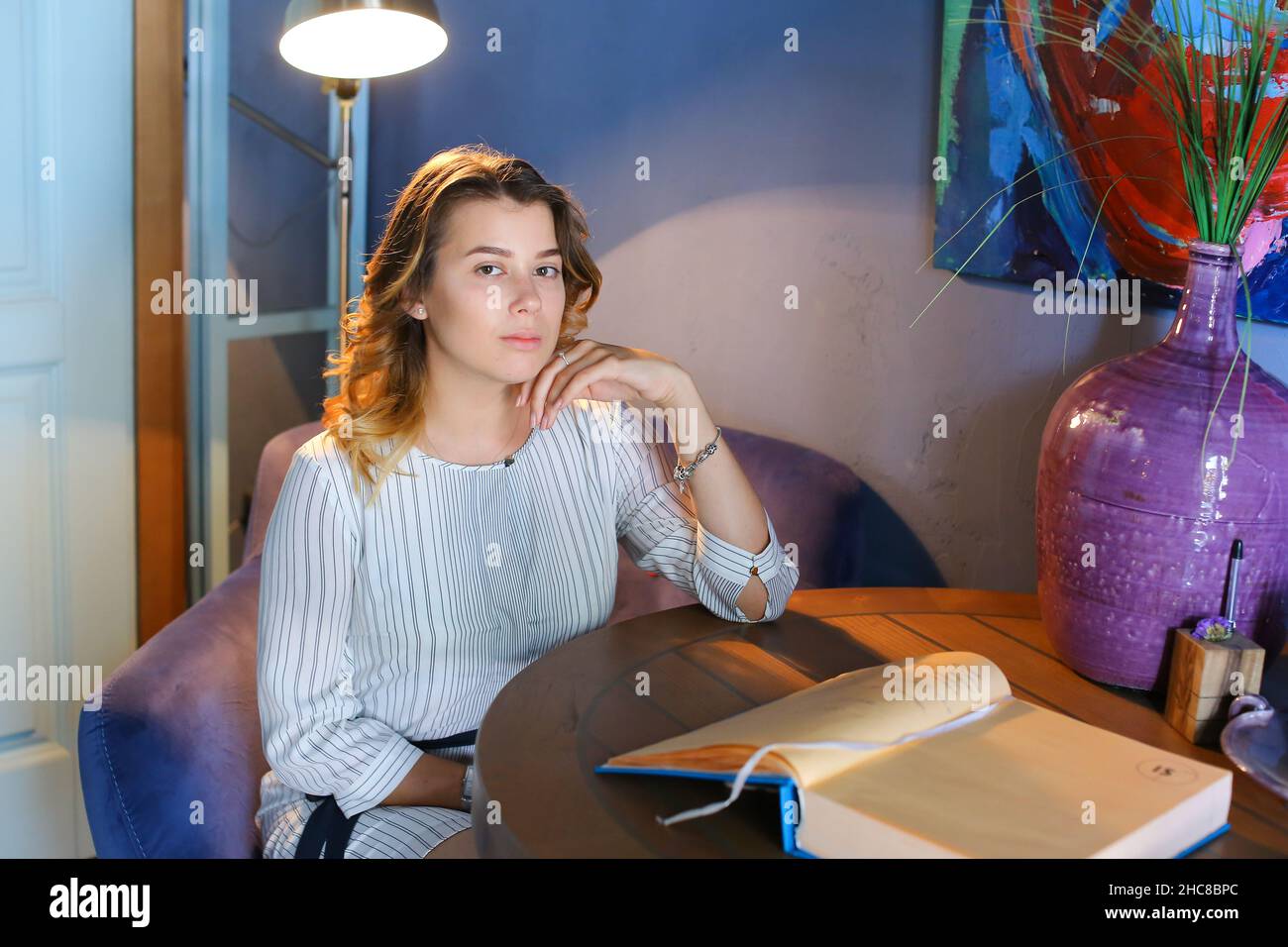Attractive young confident girl looks at camera, holds hand near face and sits in purple chair on background of lighted lamp and blue walls with Stock Photo
