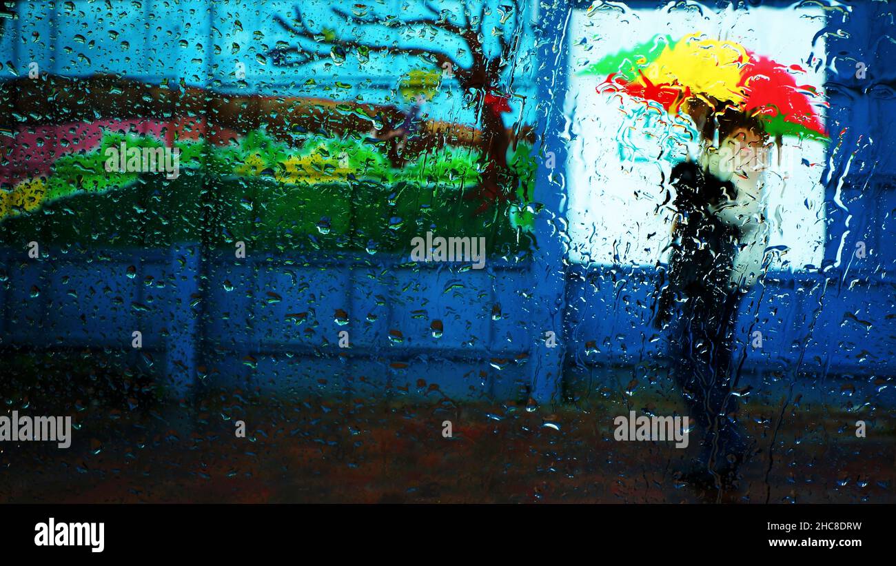 Raindrops on a glass window cause a blurred view of a winter scene of a human figure with a colourful umbrella Stock Photo