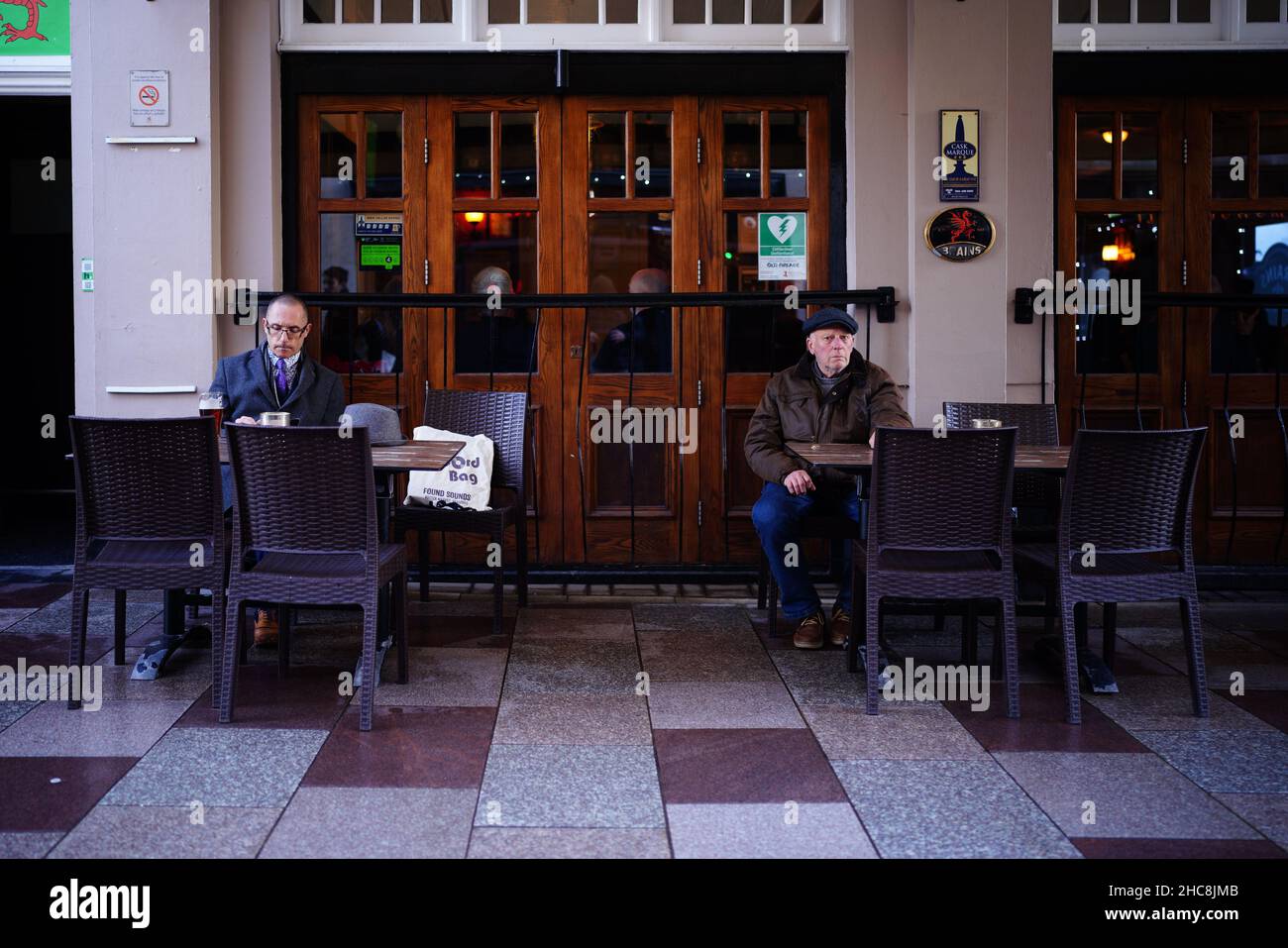 Two men sit socially distanced outside a pub in the centre of Cardiff, Wales, as new Covid-19 rules come into force. Groups of no more than six people will be allowed to meet in pubs, cinemas and restaurants in Wales, with two-metre social distancing being required in public premises and offices. Picture date: Sunday December 26, 2021. Stock Photo