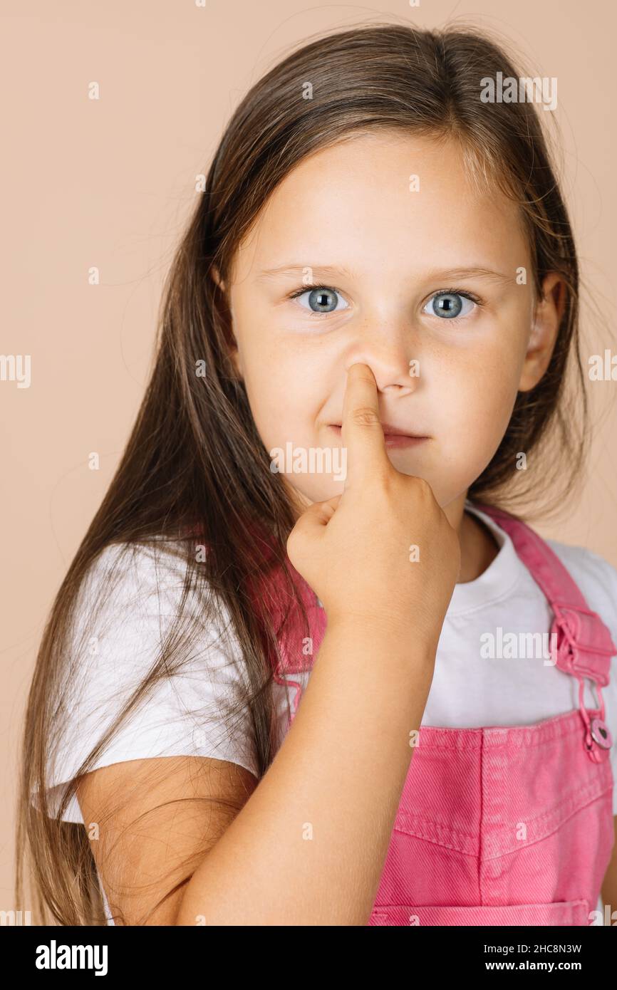 Close-up portrait female kid picking nose with point finger with shining eyes smiling looking at camera wearing bright pink jumpsuit and white t-shirt Stock Photo