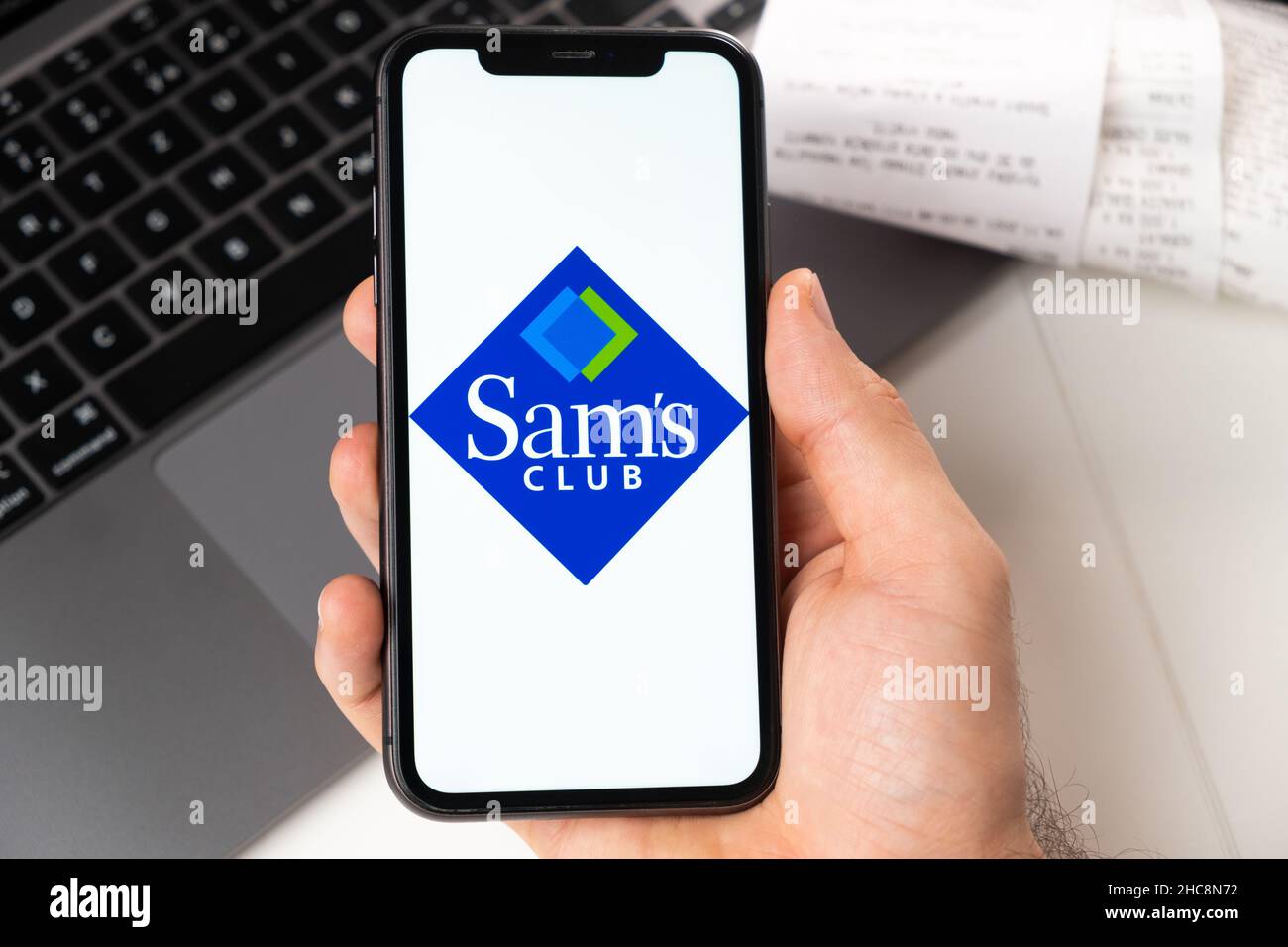 Sams Club application on the screen of Apple iPhone in mans hand and laptop on the background. Online shopping concept. November 2021, San Francisco, USA Stock Photo
