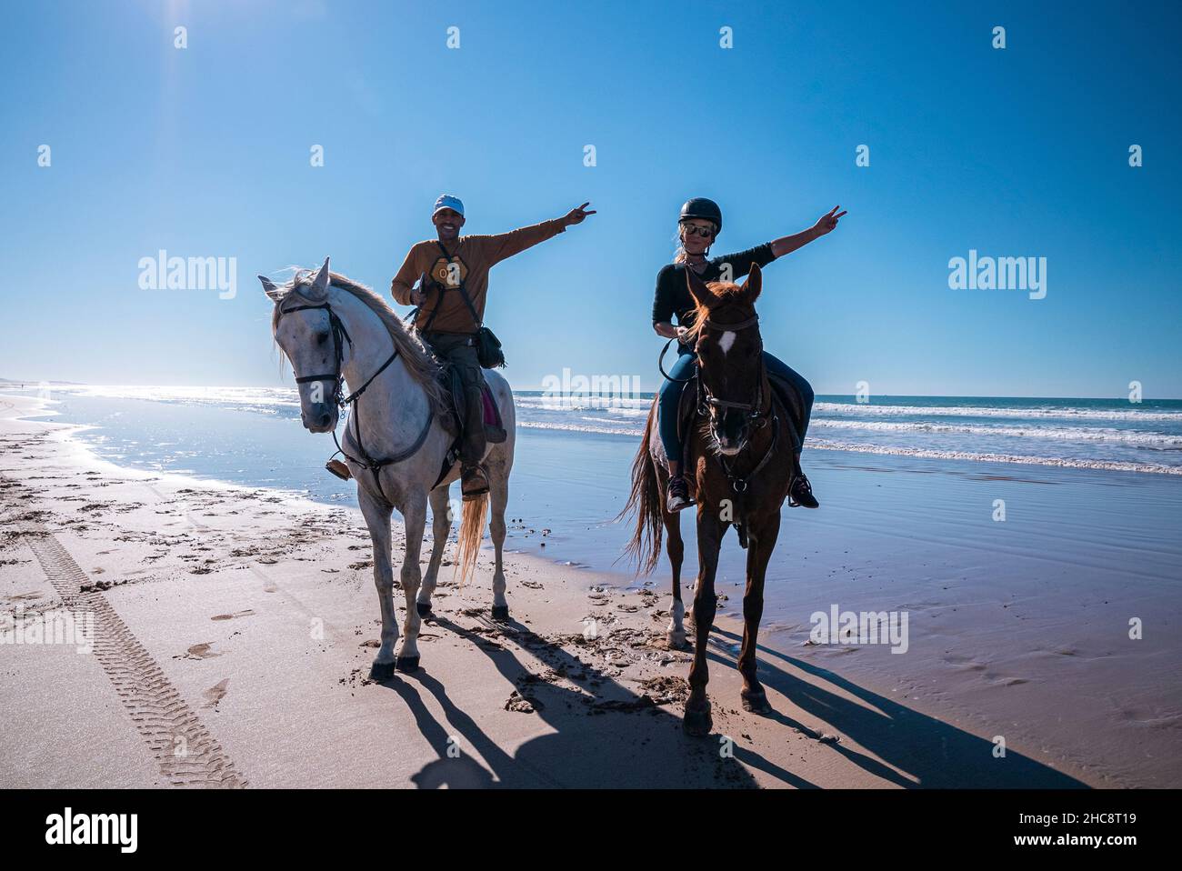 Man and woman showing victory hands gestures while sitting on horses at beach Stock Photo