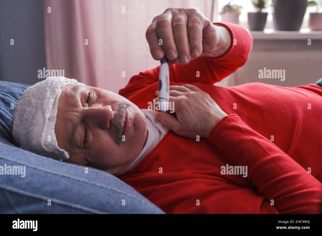 A sick male checking thermometer lying with wet towel on his forehead