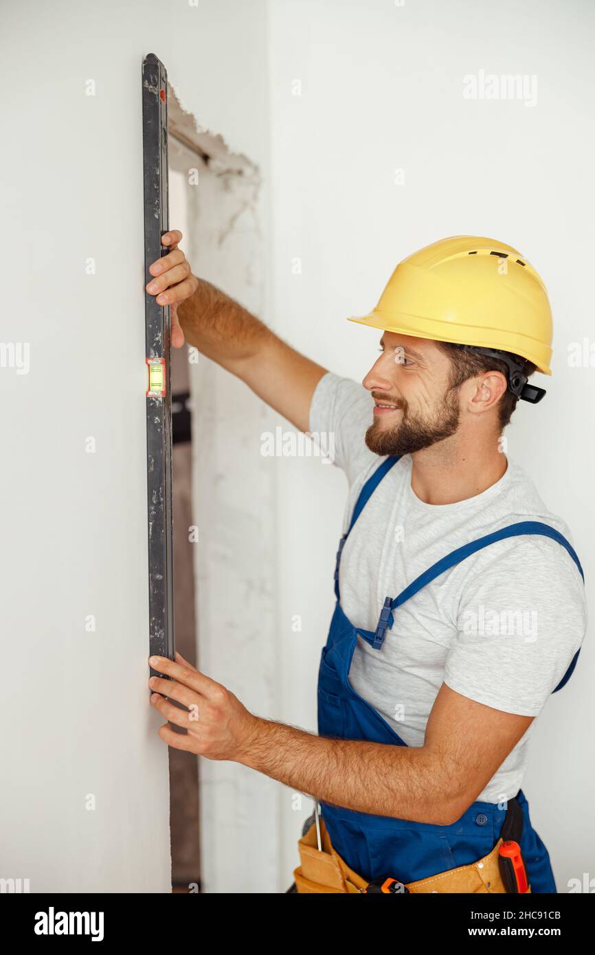 Happy handyman, builder in uniform smiling while measuring the wall using bubble level Stock Photo