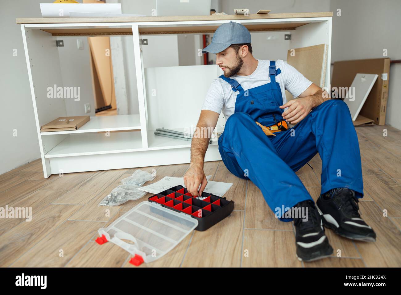 Focused handyman in overalls sitting on the floor while assembling kitchen cabinet in apartment Stock Photo