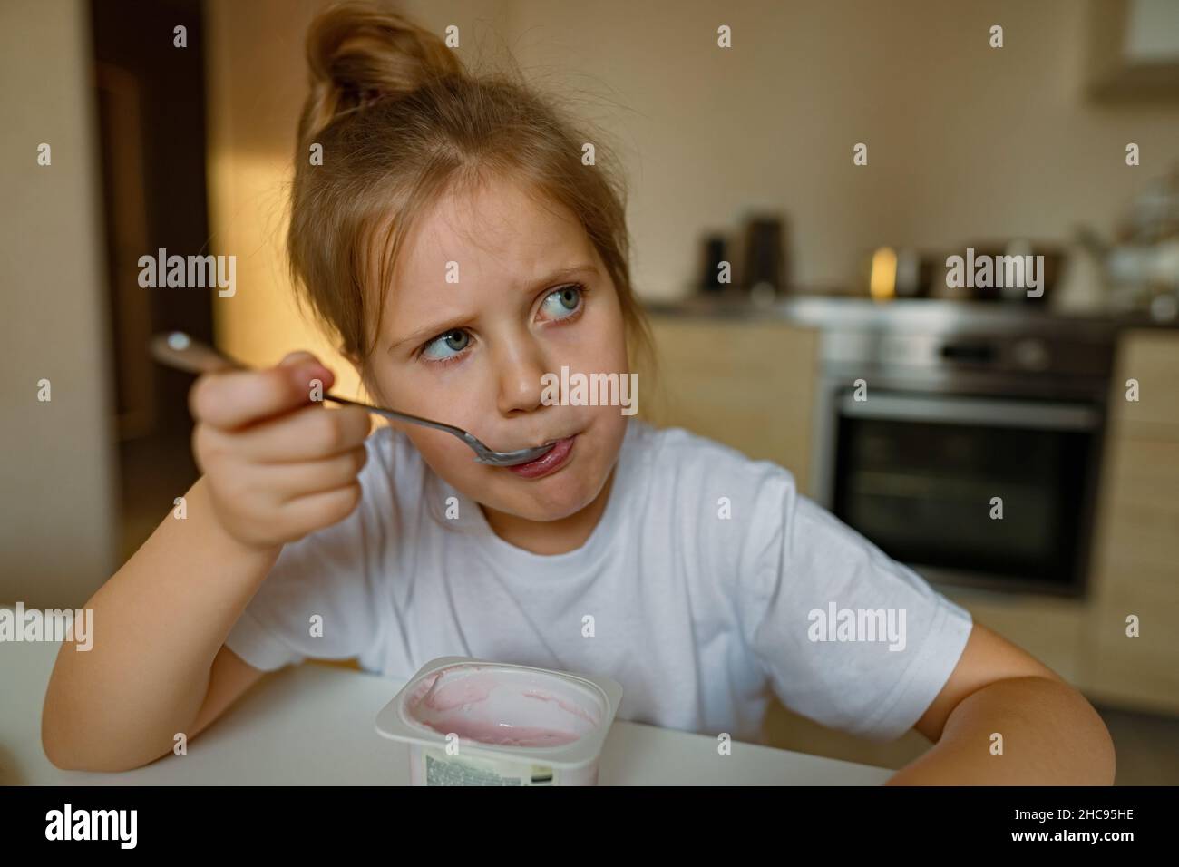 Cute girl eats yogur with appetite, has dirty mouth, has fun, grimaces Stock Photo
