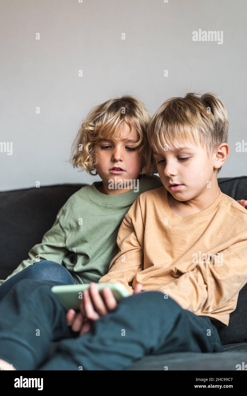 Serious little sister and brother in casual apparel with blond hair sharing mobile phone while spending weekend together sitting on couch at home Stock Photo