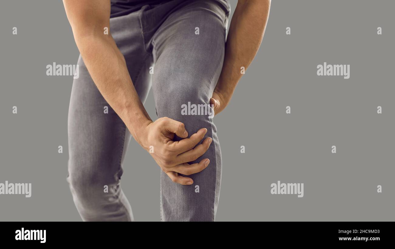 Close up of unknown man with leg trauma who is holding injured knee on gray background. Stock Photo