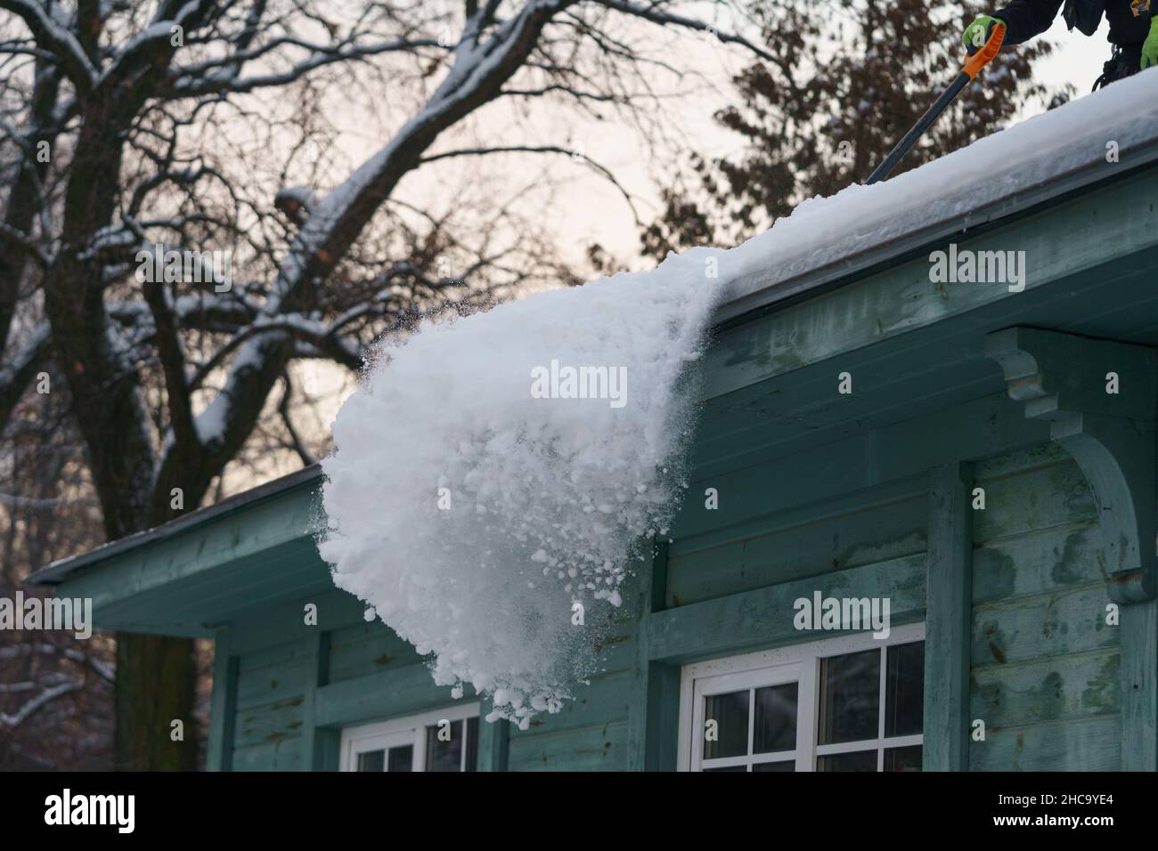 Ice dam prevention. Man holding shovel cleaning roof from snow, ice and icicles during wintertime Stock Photo