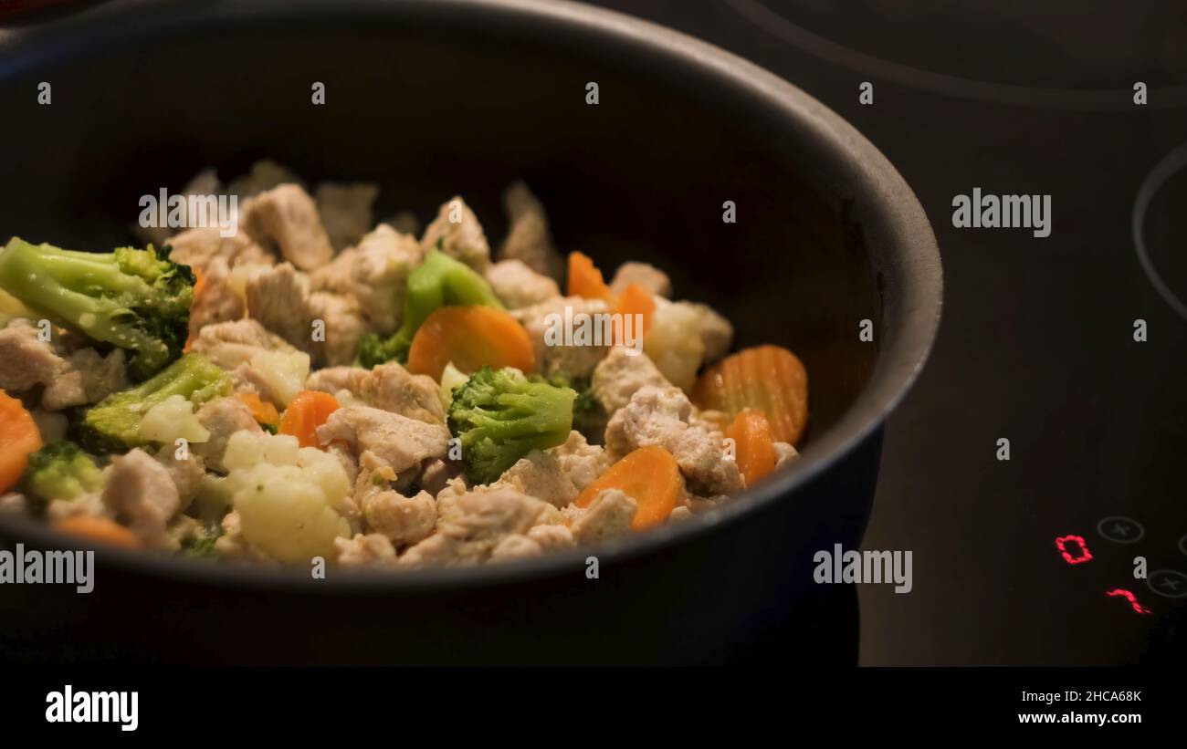 Cooking chicken with vegetables in a black frying pan standing on a touch sensitive hob. Preparing healthy dinner, frying carrots, broccoli, and cauli Stock Photo