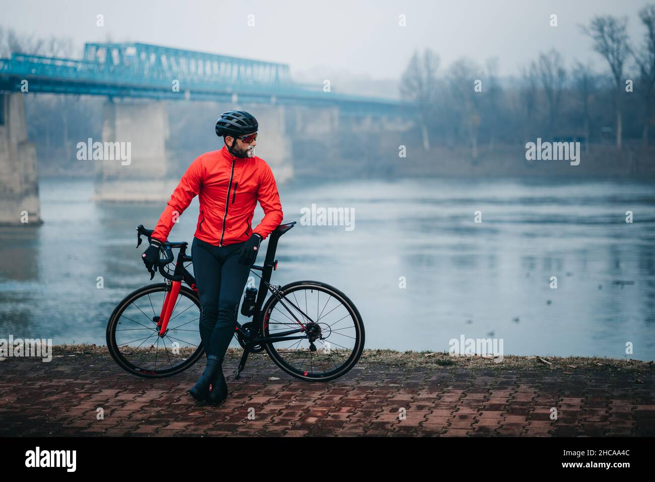 Young cool male cyclist in red sportswear posing with his bicycle by the lake on a gloomy day Stock Photo