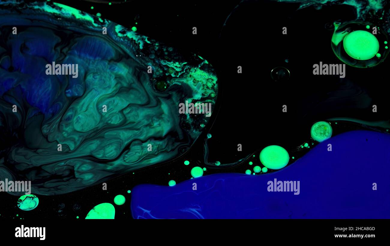 Close up of green and blue color oil paint being added into black inks. Mixed liquid paints texture on black background, art and creativity concept. Stock Photo