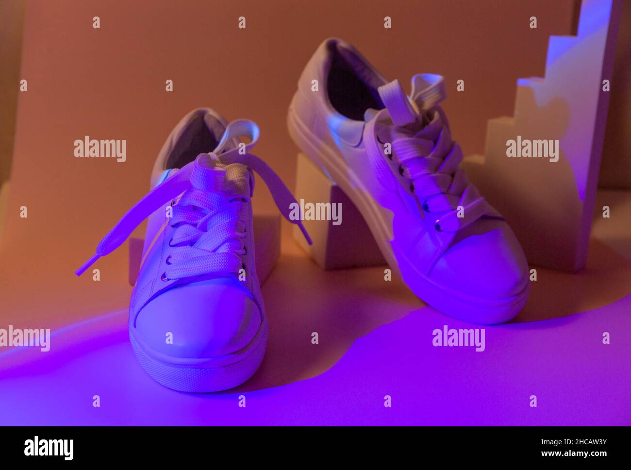Trendy fashion white sneakers on geometry podium on abstract bright background. Neon lights on casual shoes. Violet and orange gradient light. Minimal Stock Photo