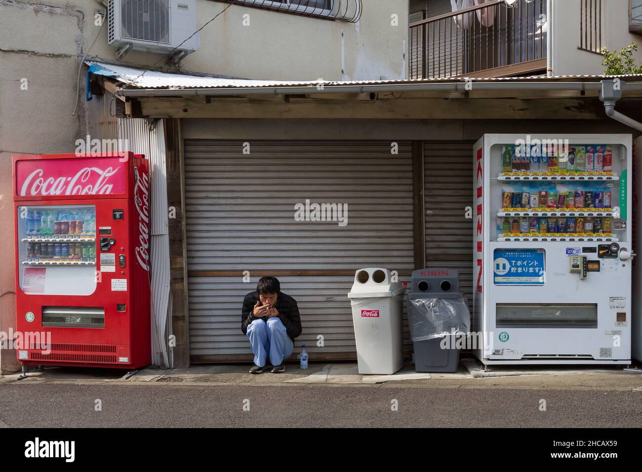 A young Japanese man squats to smoke between vending machines in Machida ward, Tokyo, Japan. Stock Photo