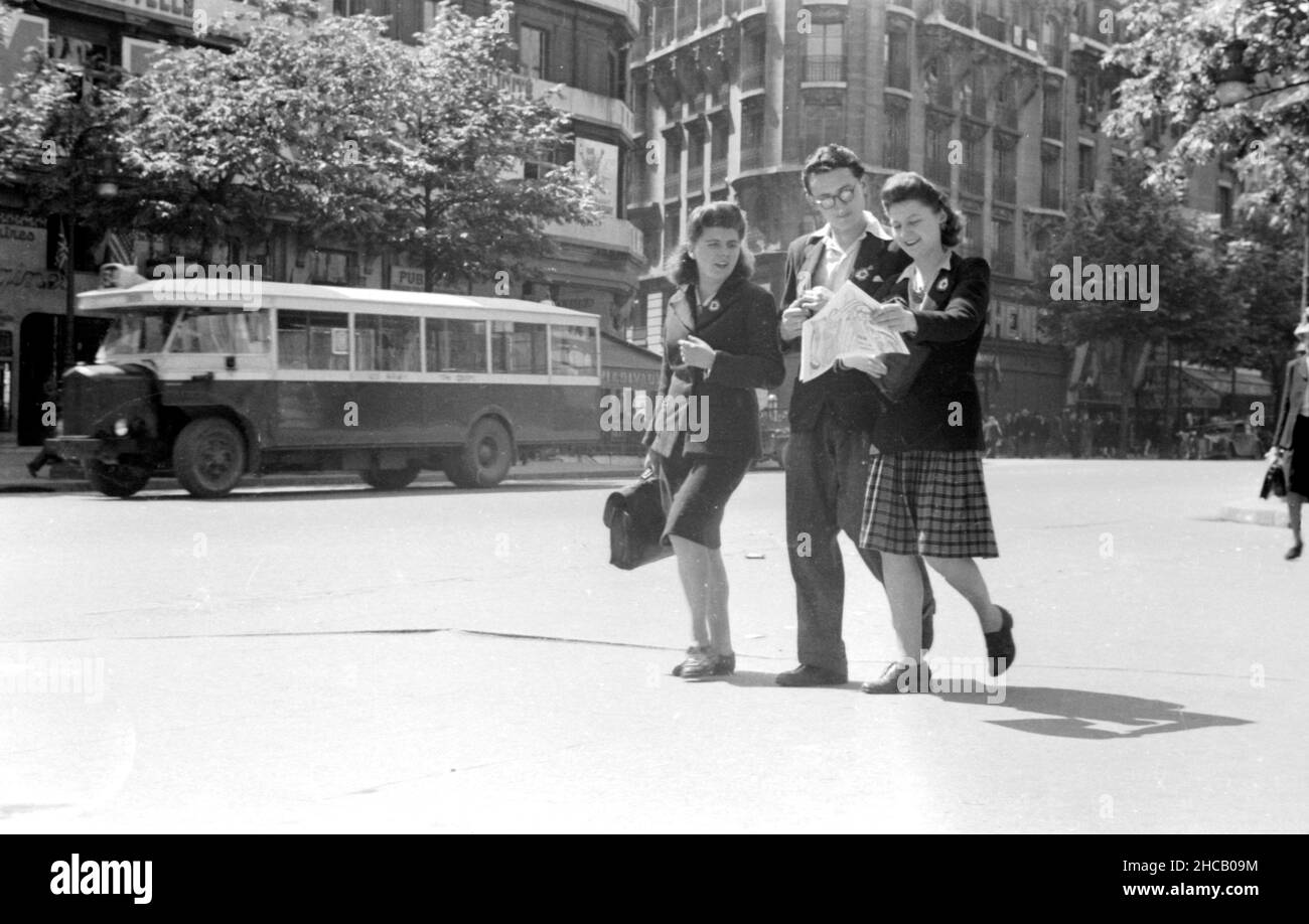 Three young people out in Paris with lapel pins, April 3 1945. They appear to be studying a circular or a map. In the background is a vintage Paris bus. This was a day of celebration in Paris that culminated with 100,000 people watching Charles De Gaulle mark the revival of the French military. De Gaulle restored colors to 131 units of the French fighting services.Swedish journalist Sven Auren reported: “Parisians were out to the man on that April morning…the place de la Concorde, where there was a veritable forest of white flagstaffs all flying the tricolour.” Stock Photo