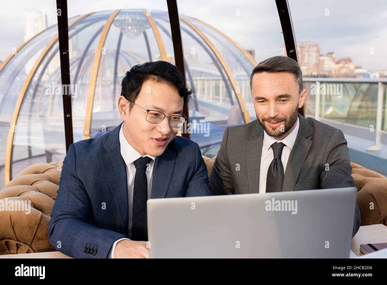 Two successful businessmen in suits looking at laptop display and discussing data while having meeting in cafe Stock Photo