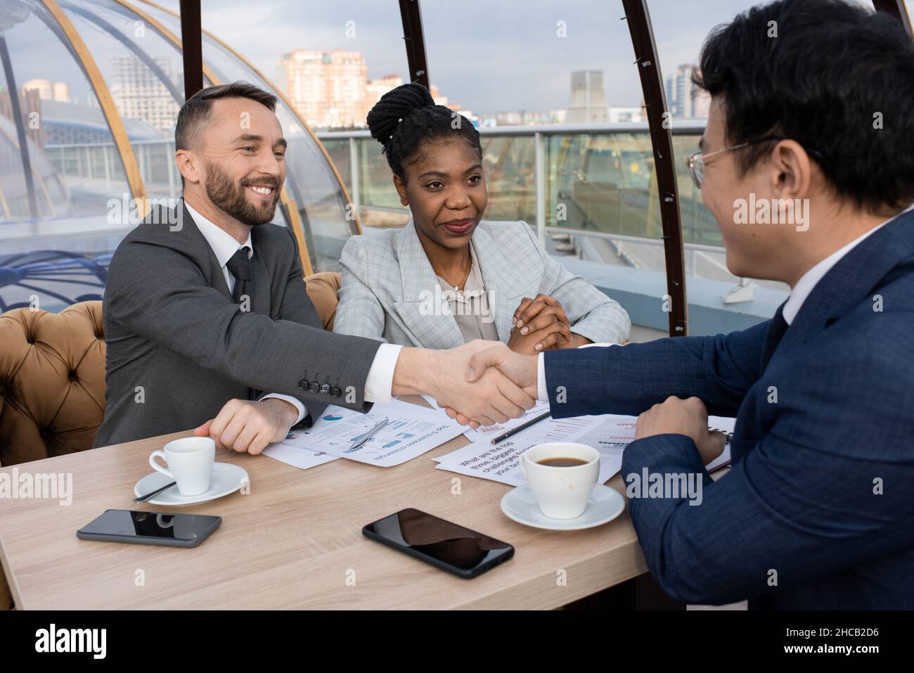 Two intercultural successful businessmen shaking hands over papers on table after negotiation about signing new contract Stock Photo