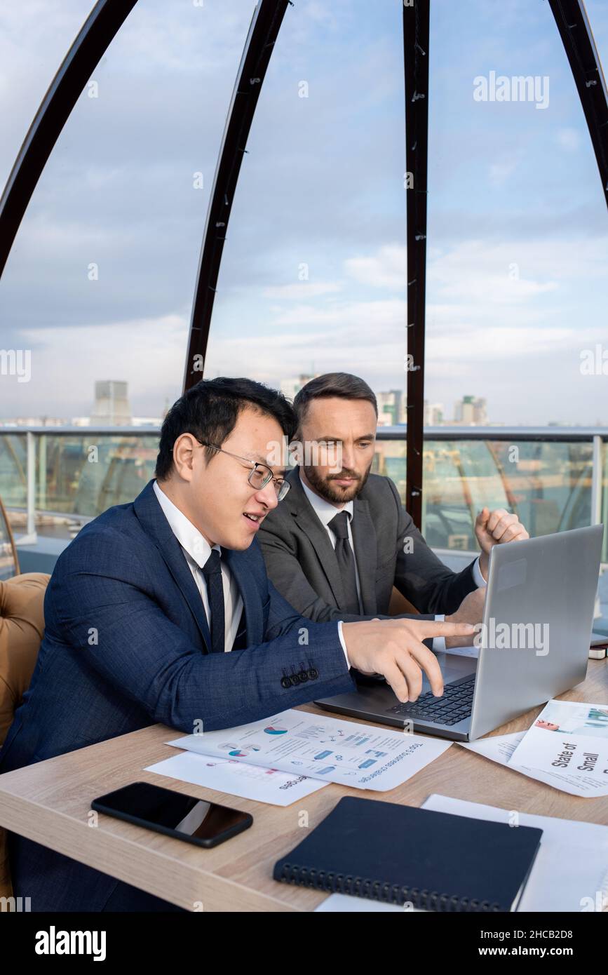 Young confident Asian businessman explaining his Caucasian colleague online data at working meeting in cafe Stock Photo
