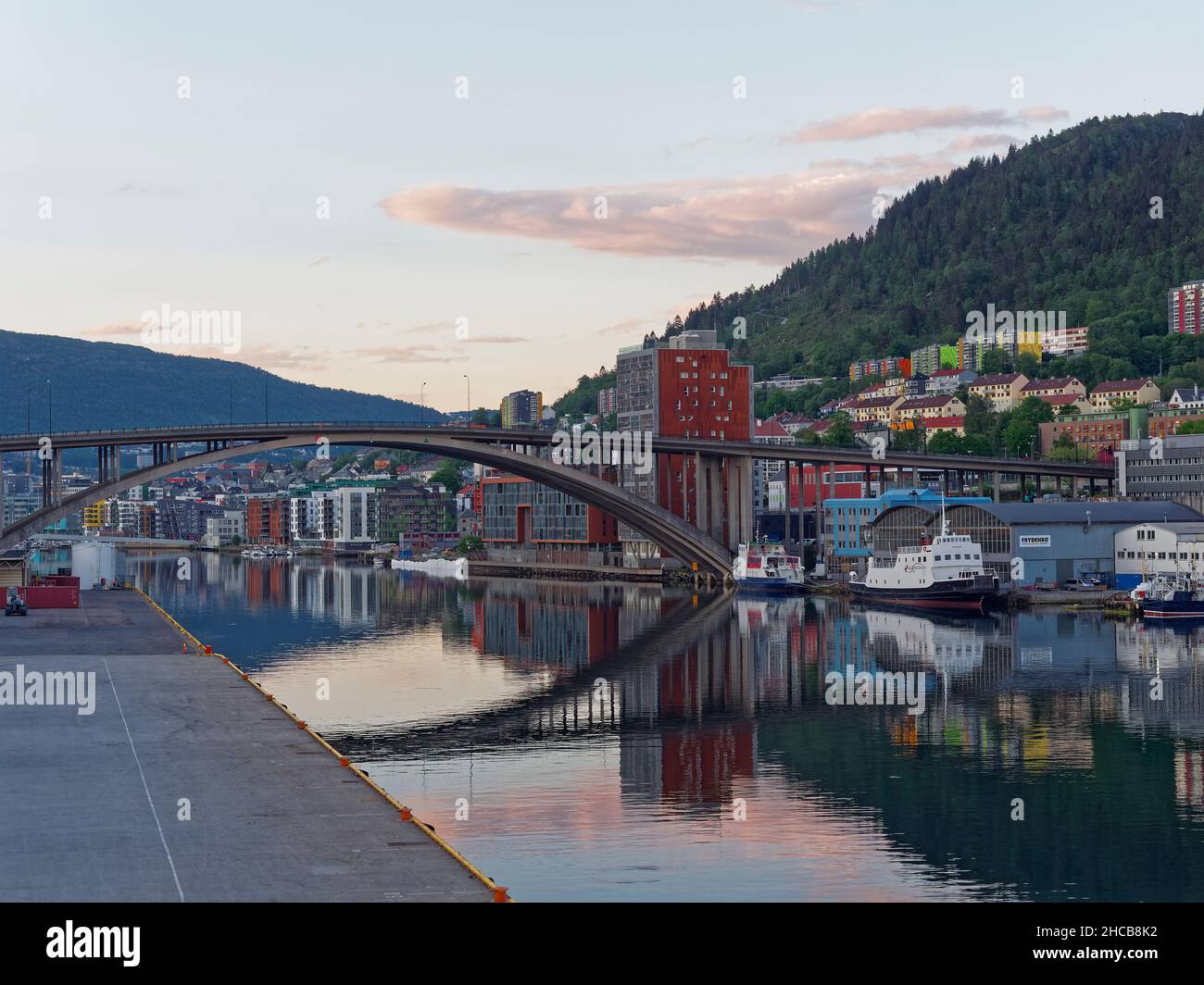 Looking through the arched road bridge and into Bergen City Centre at Dawn from the Quayside with Boats moored alongside on the calm water. Stock Photo