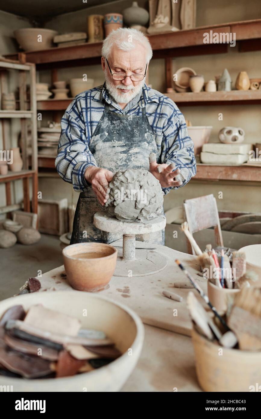 Senior man in apron using pottery wheel to make vase from clay working