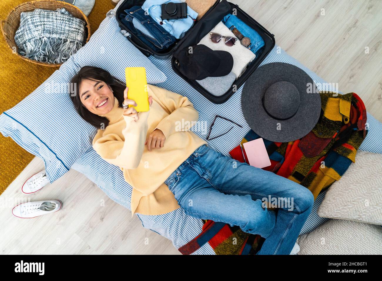 Smiling young woman taking selfie with smart phone while lying by luggage on bed Stock Photo