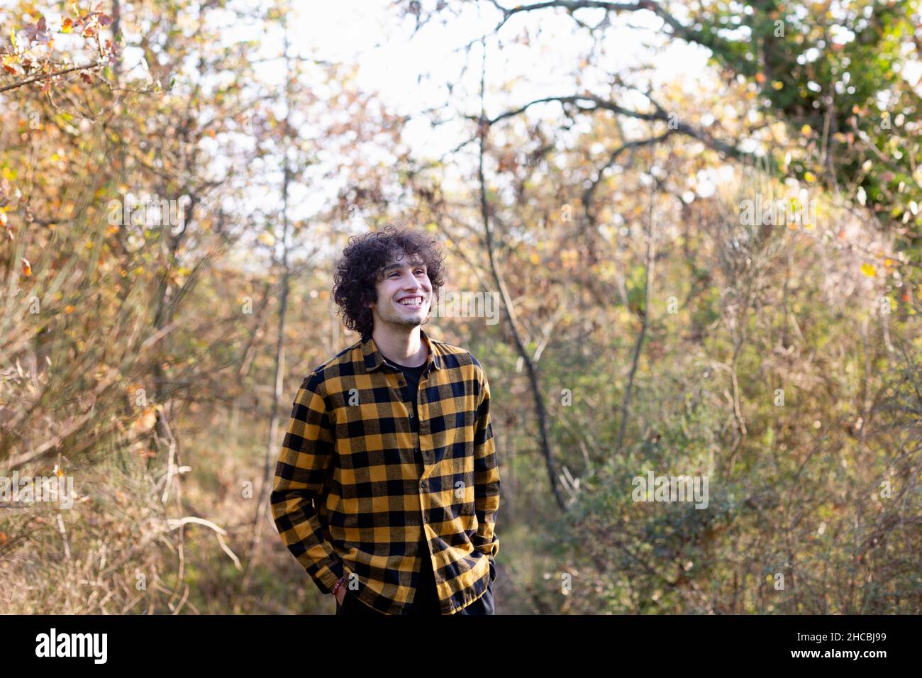 Curly haired man with hands in pockets smiling at forest Stock Photo