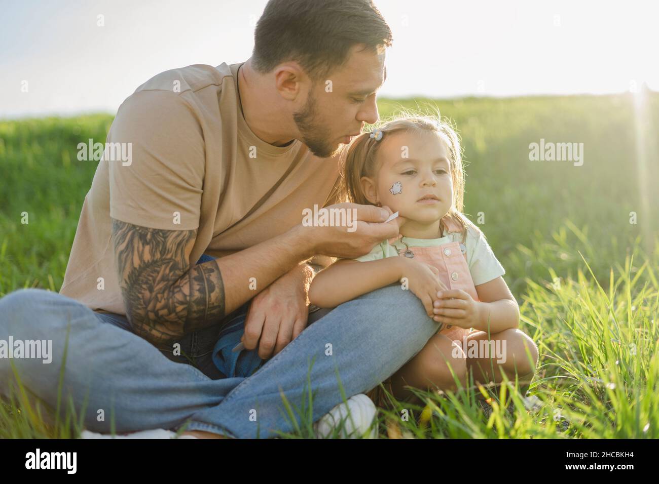 Father blowing at tattoo on daughter's face sitting on grass Stock Photo