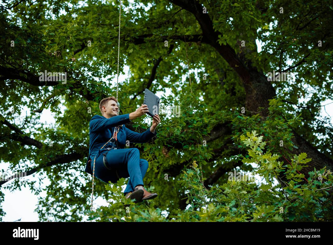 Businessman hanging on tree tied up with rope doing video call through tablet PC Stock Photo