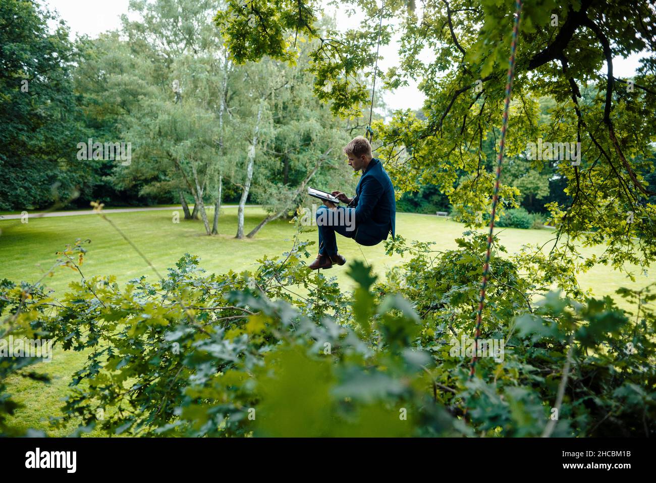 Businessman holding digital tablet tied up with rope hanging on tree Stock Photo