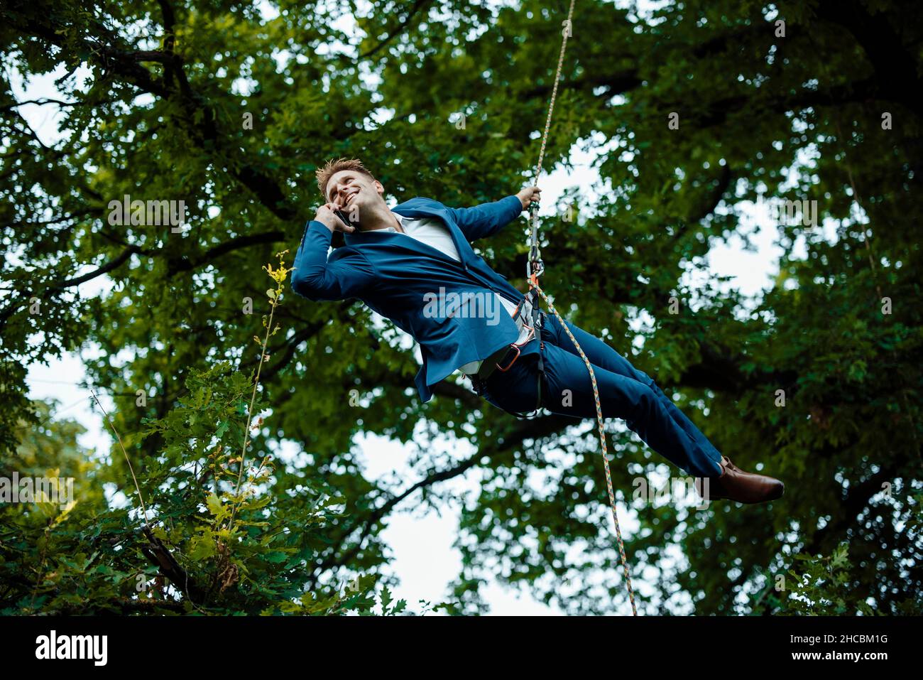 Smiling businessman hanging from rope talking on mobile phone at park Stock Photo