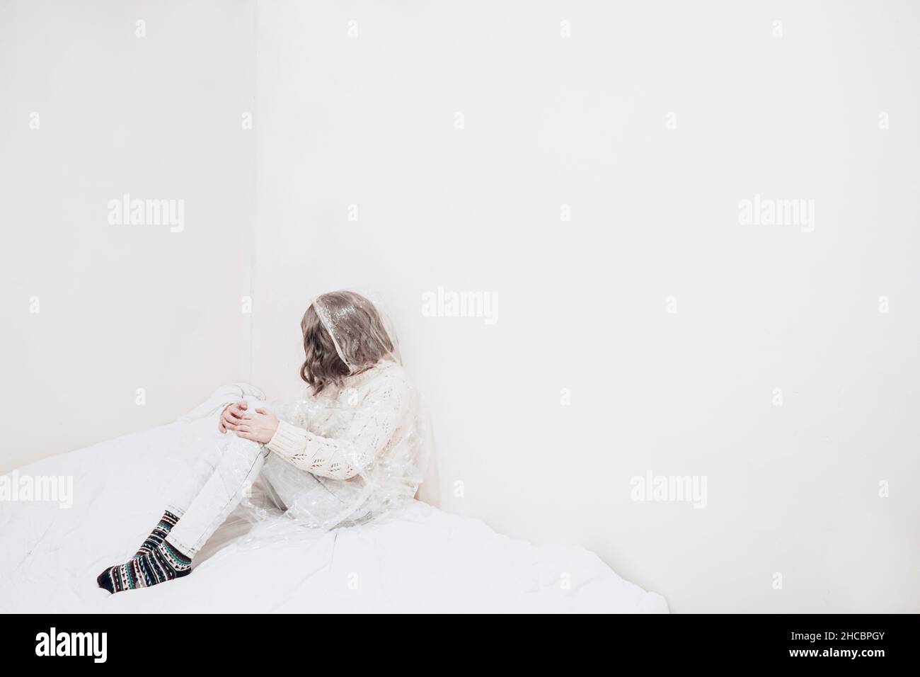 Sad woman wrapped in plastic sitting by wall on duvet Stock Photo