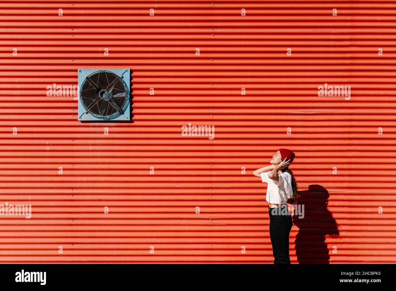 Woman wearing knit hat by red corrugated wall on sunny day Stock Photo