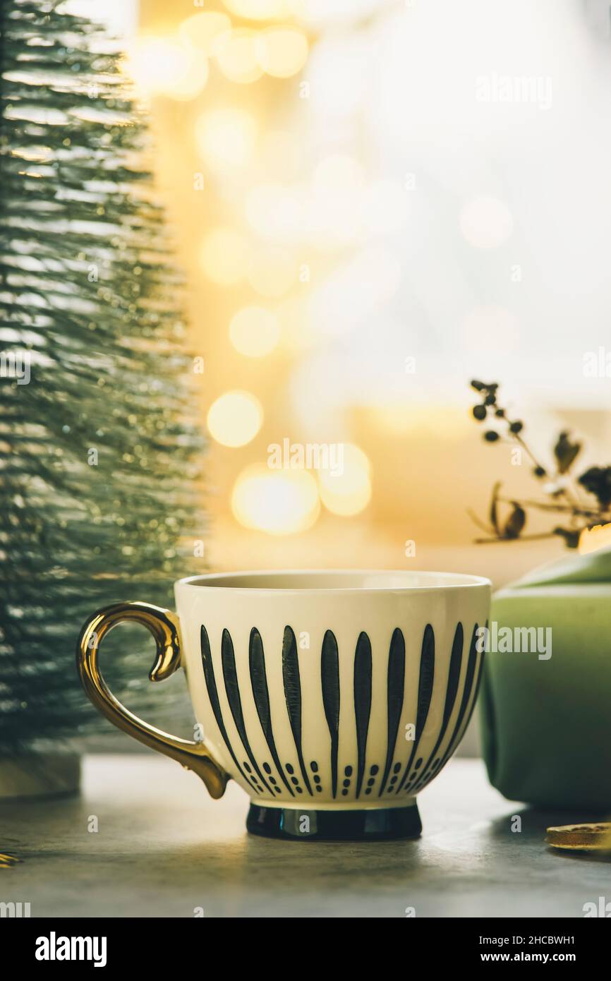 Empty striped mug on table with small Christmas tree and golden  bokeh lights. Cozy winter time at home. Front view. Stock Photo