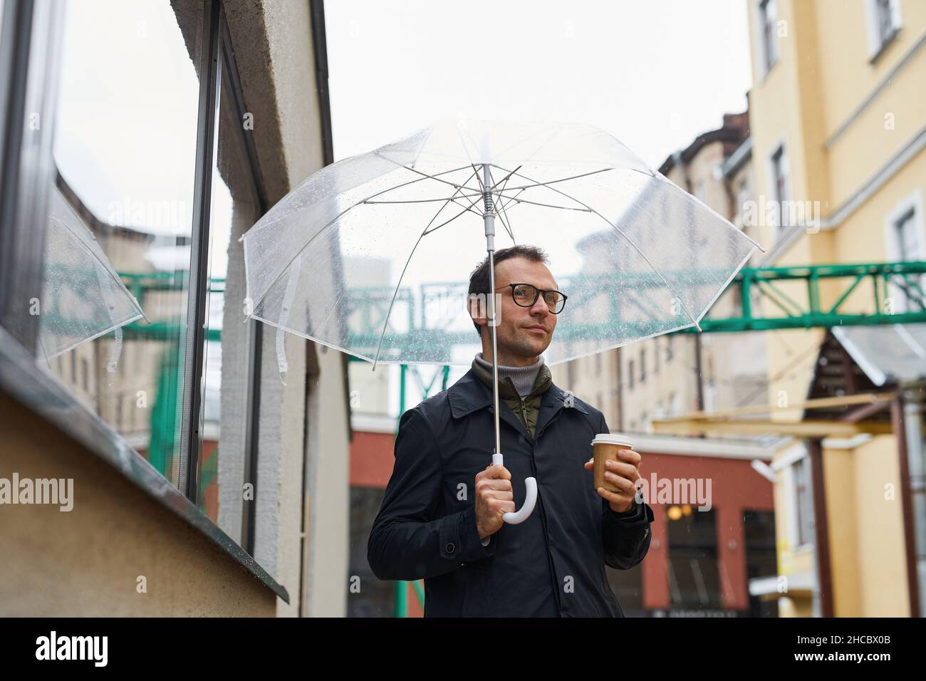 Horizontal medium shot of stylish Caucasian man wearing eyeglasses walking along street under umbrella holding coffee cup, looking away Stock Photo