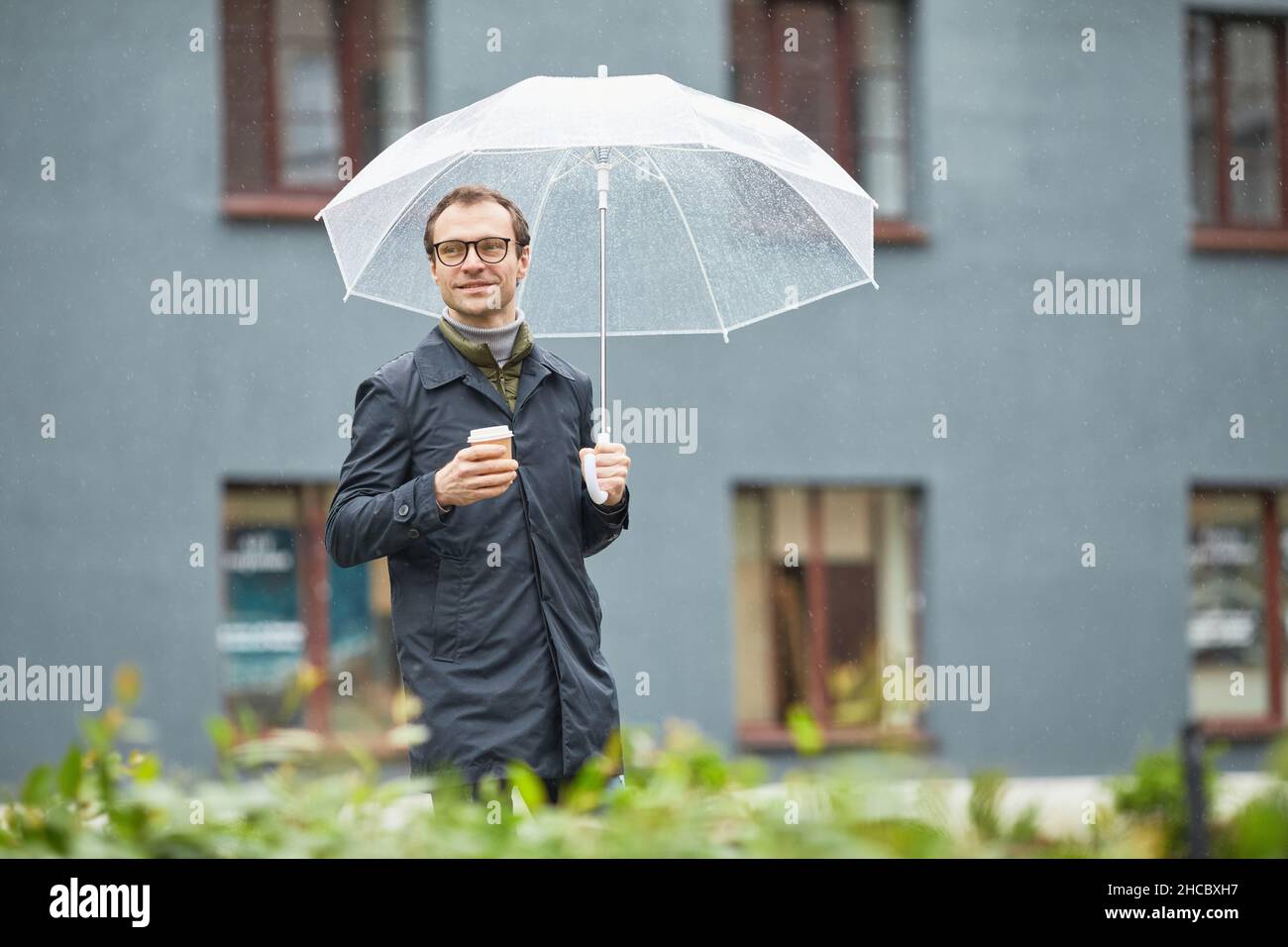 Horizontal medium shot of handsome Caucasian man wearing eyeglasses and black raincoat walking under umbrella with cup of coffee looking away Stock Photo