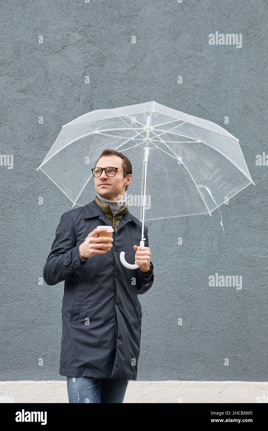 Vertical medium long shot of stylish Caucasian man wearing eyeglasses standing under umbrella with cup of coffee looking away Stock Photo