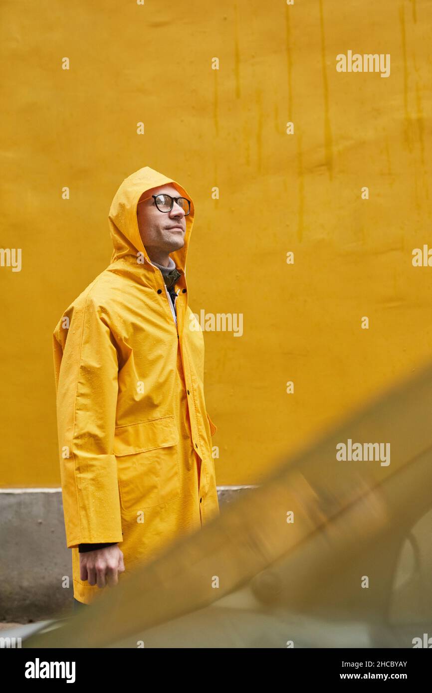 Vertical medium long shot of mature Caucasian man wearing yellow raincoat walking in rain looking away Stock Photo