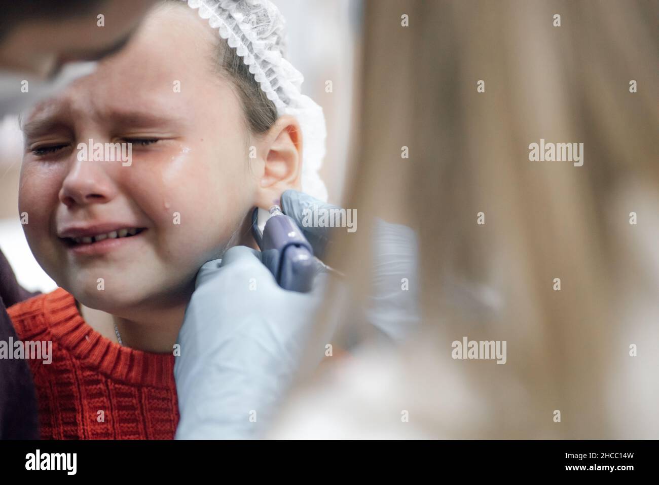 Cosmetologist wearing disposable medical gloves pierce ear with gun for piercing ears of child. Little girl crying in pain. Father support and calm Stock Photo