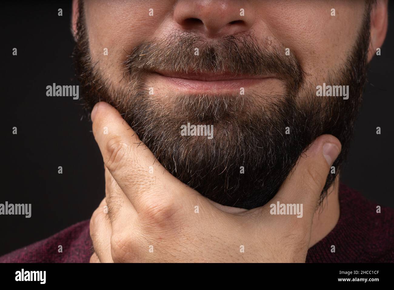 young man touches neat, trimmed black thick beard with hand after skin and hair care procedures in barbershop Stock Photo