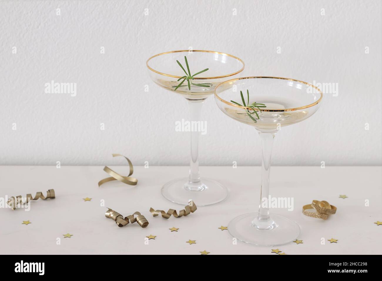 Happy New Year composition. Two champagne wine glasses with golden rim and green rosemary. White table, golden confetti stars and drinks. Party Stock Photo