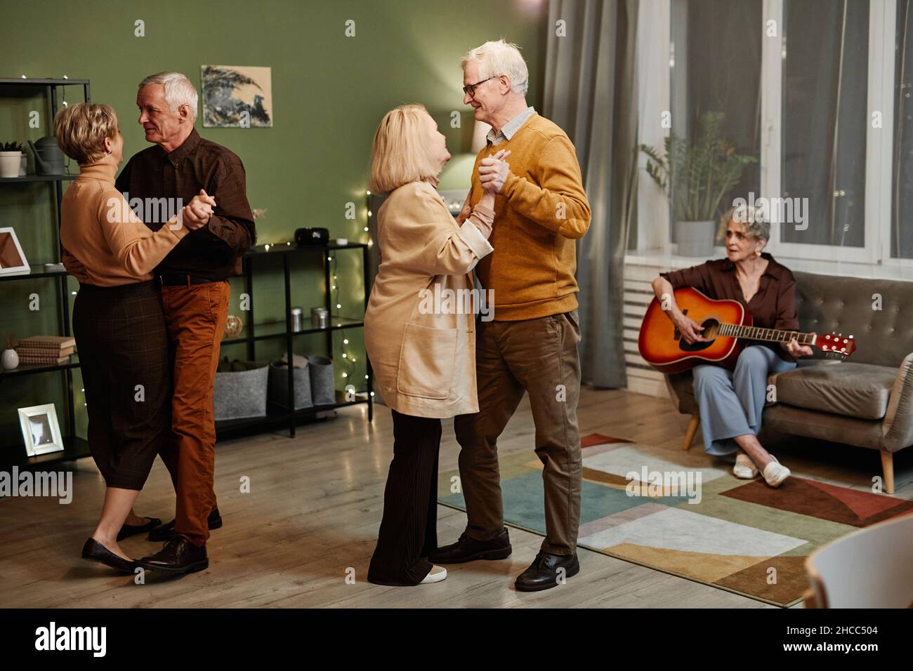 Two Caucasian senior couples dancing in decorated with lights living room while aged woman sitting on couch and playing acoustic guitar Stock Photo