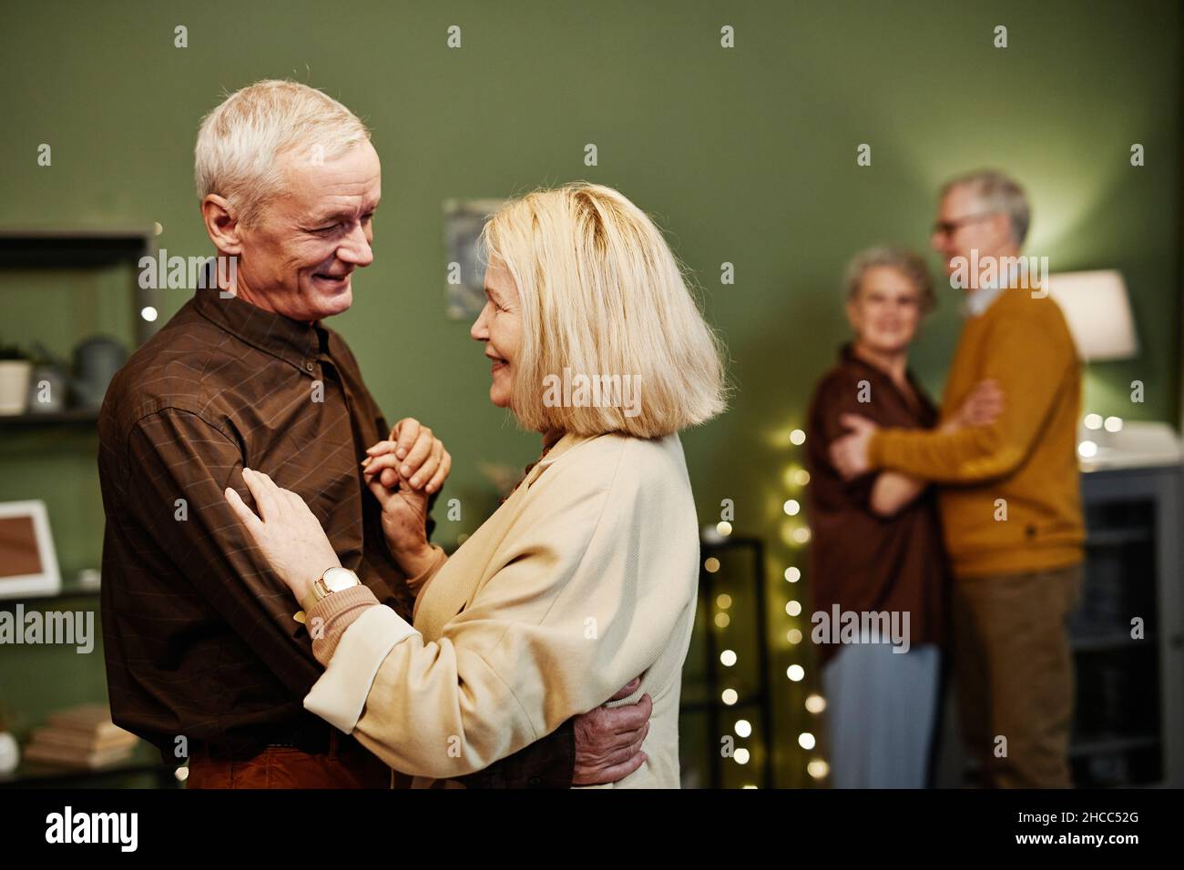 Two aged couples dancing in decorated with lights living room. Aged good-looking man with grey hair looking tenderly at his wife while dancing Stock Photo