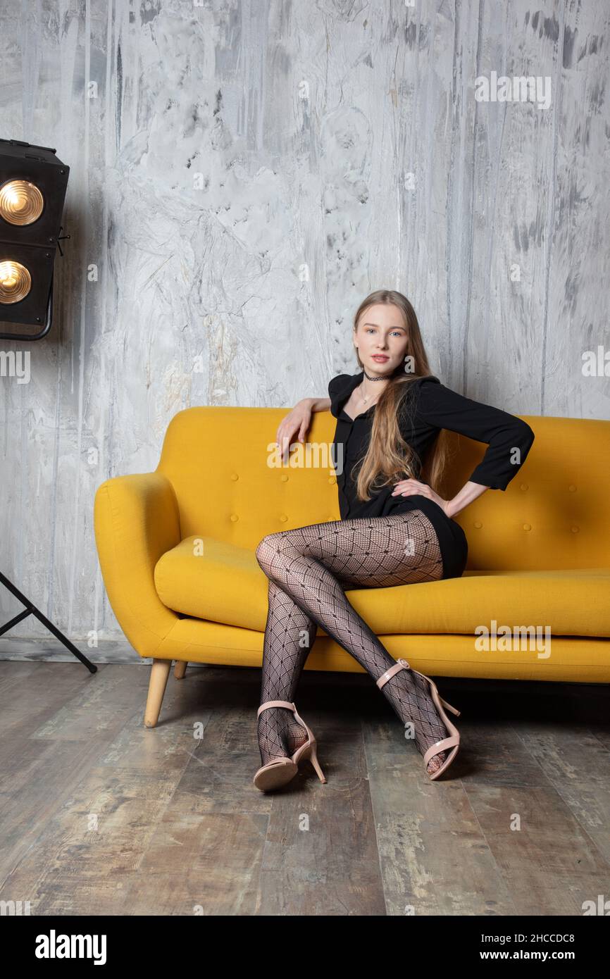 a beautiful studio shot of a stunning blonde professional model, sitting on a yellow sofa with a black short dress, high heels and tights Stock Photo