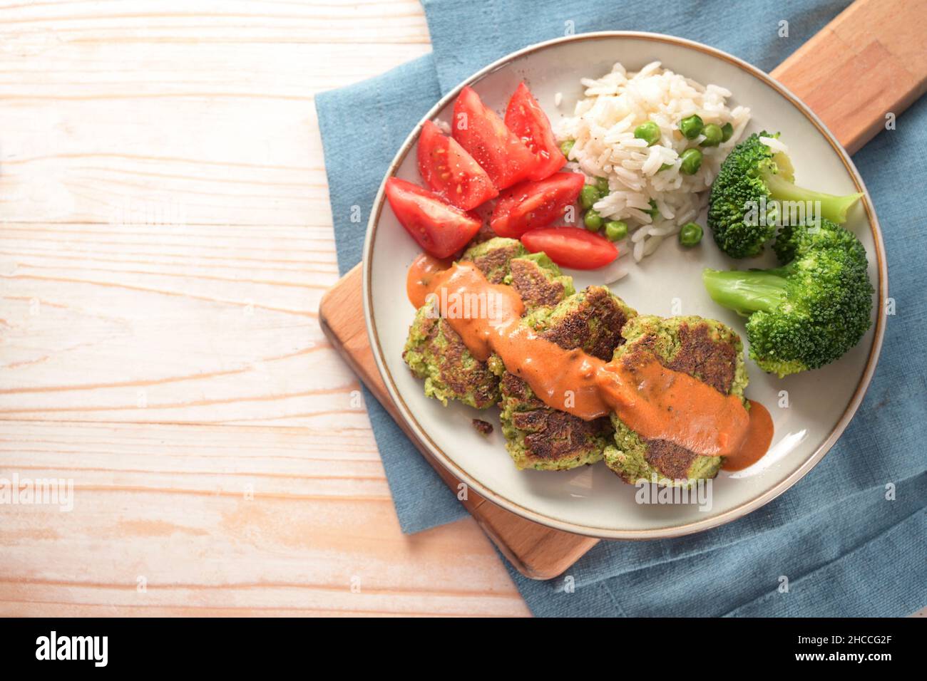 Vegetable patties with broccoli, tomato, rice and a hot sauce, vegetarian and vegan meal on a plate and a cutting board, blue napkin and light wooden Stock Photo